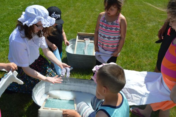 A group of children being taught how to do laundry with a washboard and soap by a women.